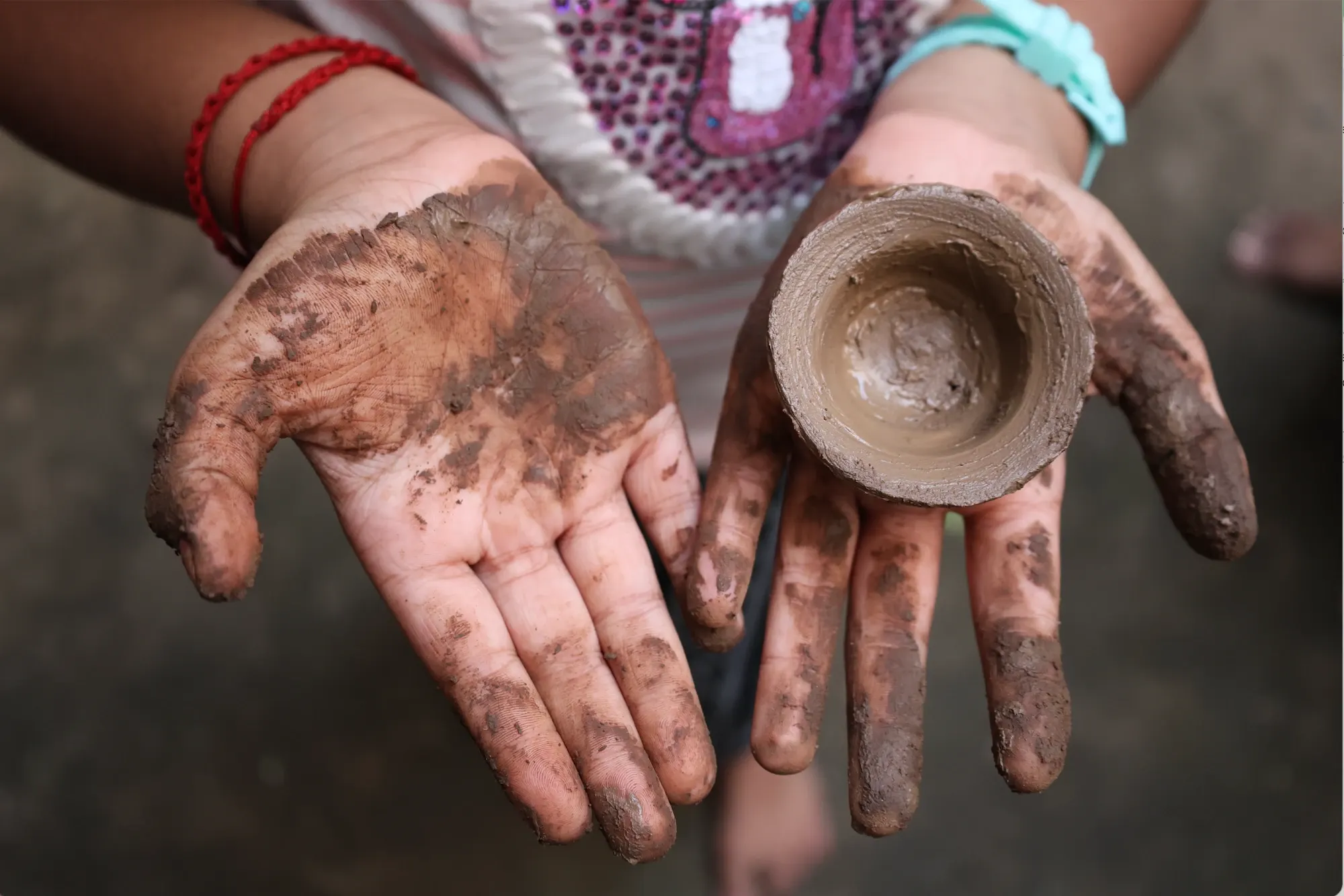 Two muddy hands hold a small clay cup made at the Sita pottery workshop